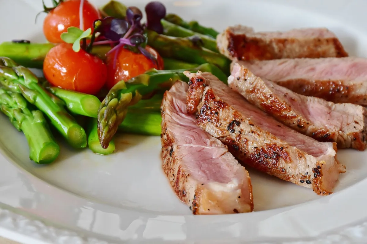 Sliced seared steak beside roasted asparagus and cherry tomatoes on a white plate.